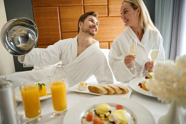 Paar genießt ein Champagner-Frühstück, während sie auf einem großen Bett sitzen - Foto, Bild