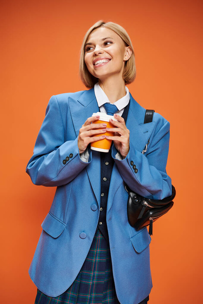 wesoła wyrafinowana kobieta z blond krótkimi włosami trzymająca torebkę i kawę na pomarańczowym tle - Zdjęcie, obraz