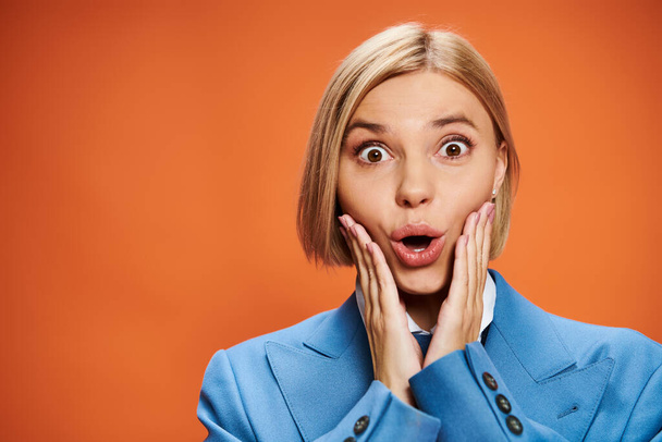 shocked young woman with short blonde hair in sophisticated attire posing on orange backdrop - Photo, Image