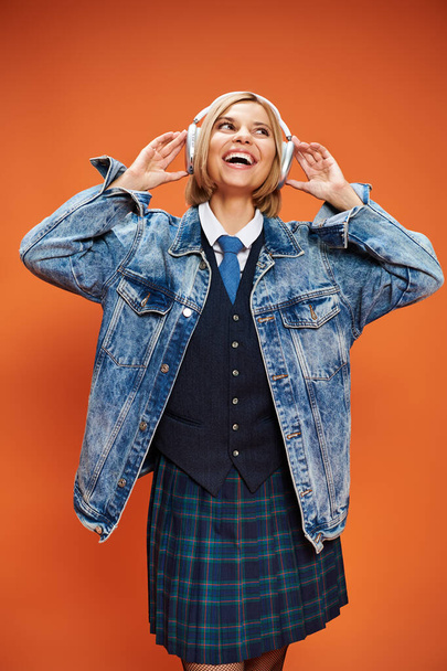 joyful stylish woman with blonde hair with headphones in denim jacket posing on orange backdrop - Photo, Image