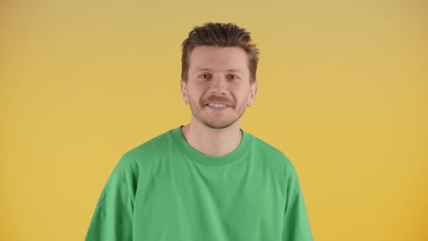 黄色の背景に隔離された緑のTシャツを着て幸せな笑顔の若い男のフロントビュー。陽気な男はカメラを見て。高品質4k映像 - 映像、動画
