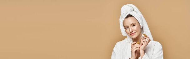 Μια φυσικά όμορφη γυναίκα κρατά παιχνιδιάρικα κρέμα, με μια πετσέτα τυλιγμένη γύρω από το κεφάλι της. - Φωτογραφία, εικόνα