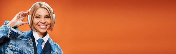 jovem alegre com cabelo loiro com fones de ouvido em jaqueta jeans posando no fundo laranja, banner - Foto, Imagem