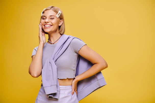 joyous well dressed woman with blonde short hair in vibrant attire posing on yellow backdrop - Photo, Image