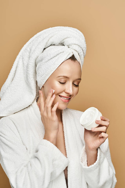 Uma mulher bonita com uma toalha enrolada em torno de sua cabeça segura um frasco de creme, exalando beleza natural e elegância. - Foto, Imagem
