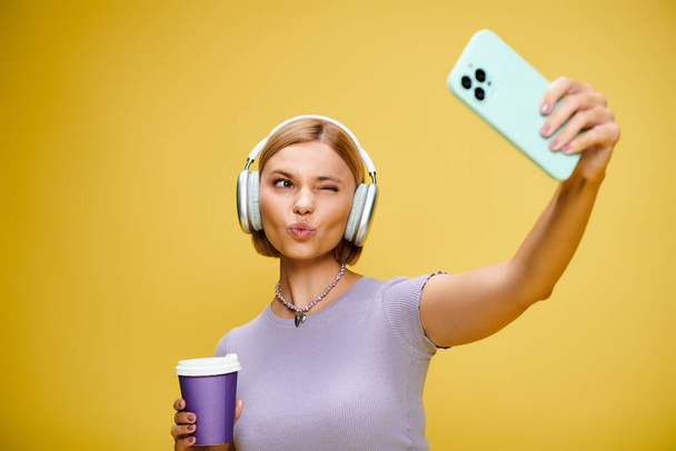séduisante femme gaie avec écouteurs et téléphone portable profitant de son café sur fond jaune - Photo, image