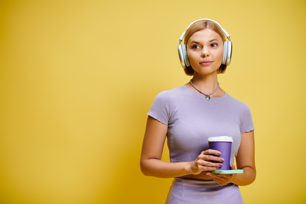 junge, fröhliche Frau mit Kopfhörer und Handy genießt ihren Kaffee vor gelbem Hintergrund - Foto, Bild