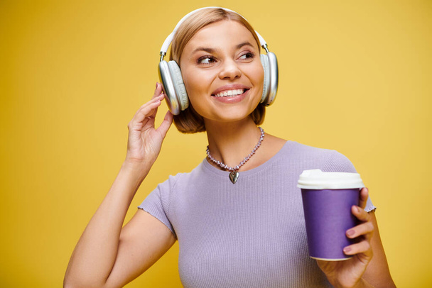 весела стильна жінка з навушниками насолоджується музикою і гарячою кавою, позуючи на жовтому тлі - Фото, зображення