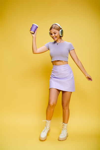 iloinen trendikäs nainen kuulokkeet nauttia musiikkia ja kuumaa kahvia poseeratessaan keltainen tausta - Valokuva, kuva