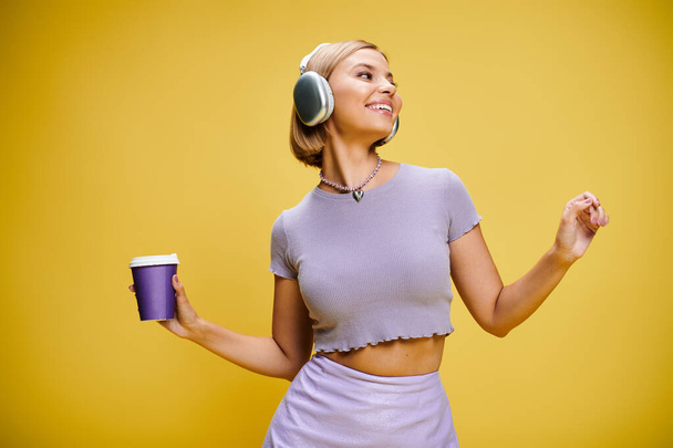 alegre mujer de moda con auriculares disfrutando de la música y el café caliente mientras posa en el fondo amarillo - Foto, imagen