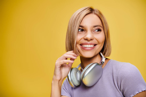 Charmante, fröhliche Frau mit kurzen blonden Haaren und Kopfhörern, die Musik auf gelbem Hintergrund genießt - Foto, Bild