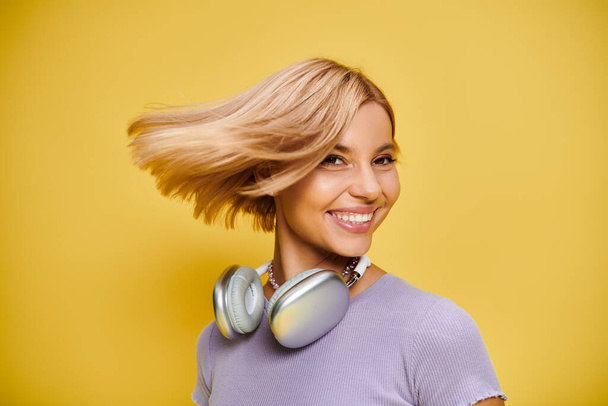 黄色い背景で音楽を楽しんでいる短いブロンドの髪とヘッドフォンの洗練された喜びの女性 - 写真・画像