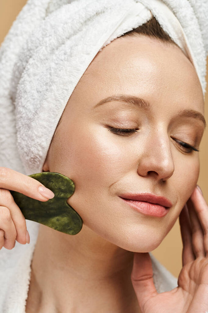 Kobieta z ręcznikiem owiniętym wokół głowy pozuje z zieloną gua sha na twarzy, pokazując swoje naturalne piękno. - Zdjęcie, obraz