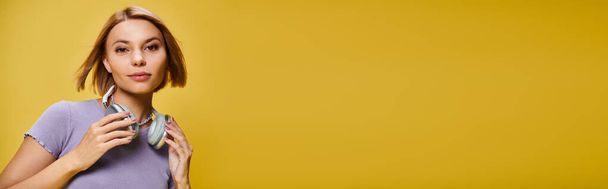stylowa radosna kobieta z krótkimi blond włosami i słuchawkami ciesząca się muzyką na żółtym tle, baner - Zdjęcie, obraz
