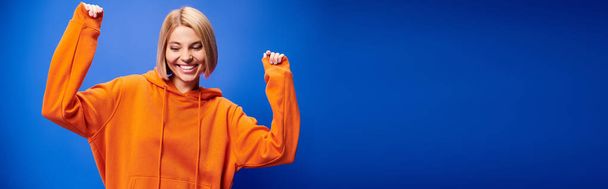 tevreden vrouw met kort haar in levendige oranje hoodie poseren actief op blauwe achtergrond, banner - Foto, afbeelding