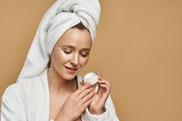 Piękna kobieta z ręcznikiem na głowie trzyma słoik śmietany, pokazując jej naturalne piękno i rutynę pielęgnacji skóry. - Zdjęcie, obraz