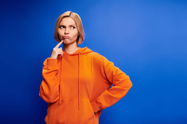 alluring puzzled blonde woman with short hair in stylish orange hoodie posing on blue background - Photo, Image