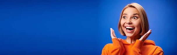 positive Frau mit kurzen Haaren in leuchtend orangefarbenem Kapuzenpulli posiert aktiv vor blauem Hintergrund, Banner - Foto, Bild