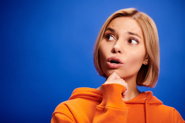 appealing puzzled blonde woman with short hair in stylish orange hoodie posing on blue background - Photo, Image