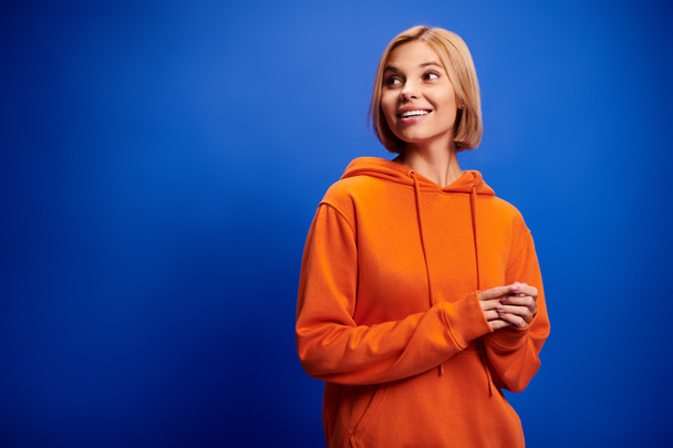 appealing cheerful woman with short hair in vibrant orange hoodie posing actively on blue backdrop - Photo, Image