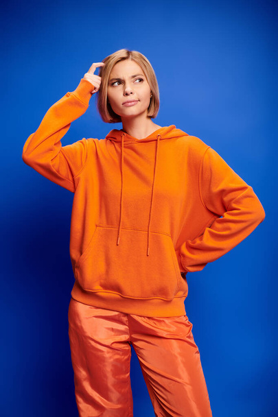 confusa mujer rubia atractiva con pelo corto en sudadera con capucha naranja elegante posando sobre fondo azul - Foto, Imagen