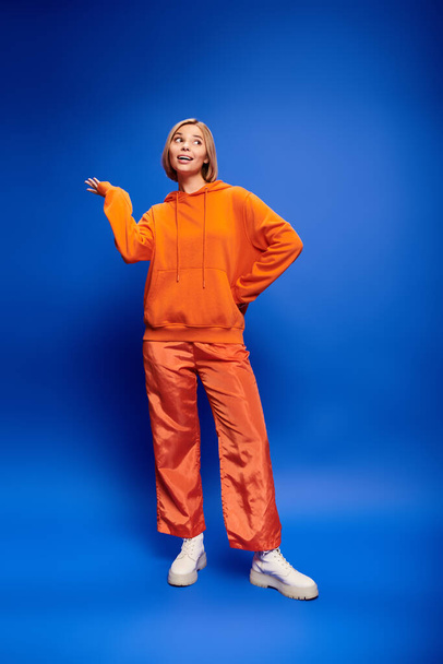 alluring cheerful woman with short hair in vibrant orange hoodie posing actively on blue backdrop - Photo, Image