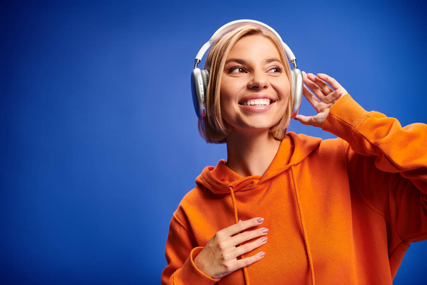 привлекательная веселая женщина с короткими светлыми волосами и наушниками, наслаждающаяся музыкой на синем фоне - Фото, изображение