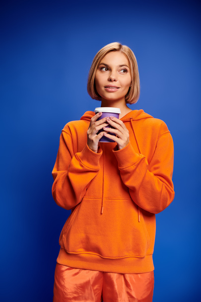 attraktive fröhliche Frau mit kurzen blonden Haaren in lebendiger Kleidung mit Kaffeetasse vor blauem Hintergrund - Foto, Bild