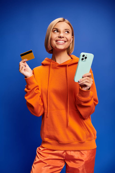 bela mulher alegre com cabelo loiro com capuz laranja vibrante posando com cartão de crédito e telefone - Foto, Imagem