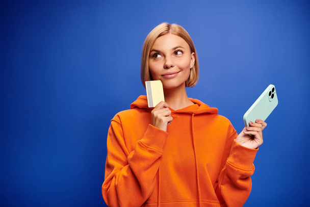 atrakcyjna radosna kobieta z blond włosami w pomarańczowym kapturze pozująca z kartą kredytową i telefonem - Zdjęcie, obraz
