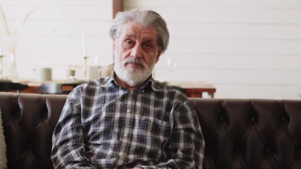 Un uomo dai capelli grigi si siede sul divano del soggiorno e guarda direttamente la telecamera. Filmati 4k di alta qualità - Filmati, video