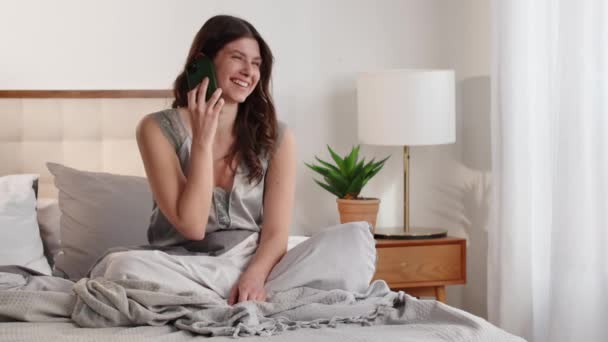 Nuori nainen istuu sängyllä ikkunan vieressä ja puhuu puhelimessa. Hymyilevä nainen keskustelee puhelimessa ystävänsä kanssa tulevan kävelyn yksityiskohdista ympäri kaupunkia. Korkea laatu - Materiaali, video