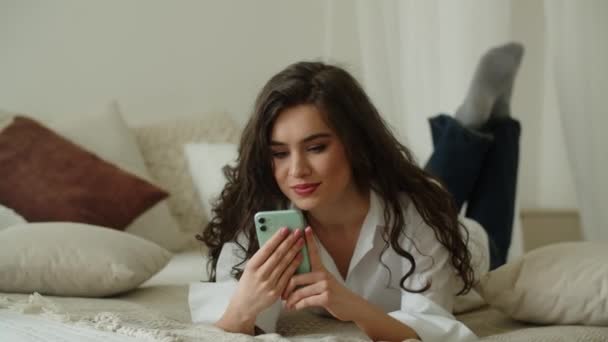 Chica riendo leyendo mensaje de teléfono mientras está acostada en la cama en su habitación. Mujer feliz con smartphone. Buenas noticias. Imágenes de alta calidad 4k - Metraje, vídeo