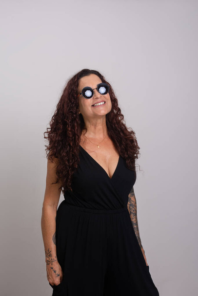 Красива кучерява жінка в сонцезахисних окулярах і чорному одязі посміхається. Ізольовані на білому тлі. - Фото, зображення