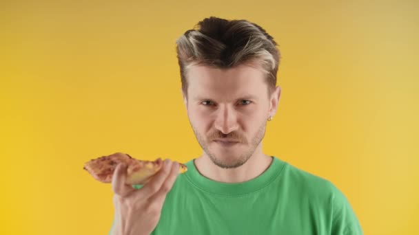 若い男がカメラを覗き込み、ピザを噛んだ。黄色い背景のカメラの前には緑色のTシャツを着た男が立っていて、ピザを食べている。高品質4k映像 - 映像、動画