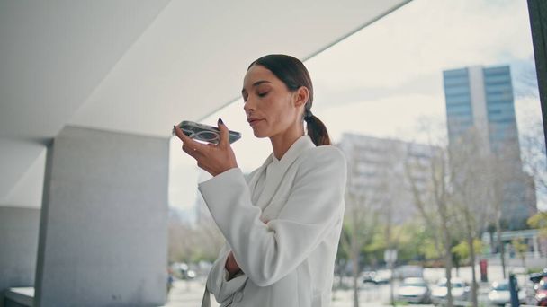 Businesswoman using voice recognition virtual assistant at phone standing at modern building close up. Confident serious woman recording audio message on telephone. Rich lady speaking in loudspeaker. - Photo, Image