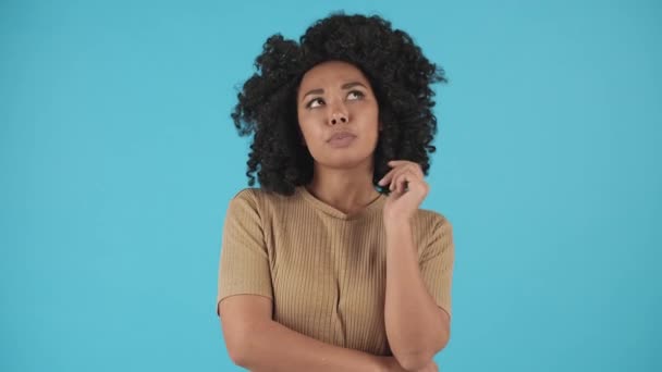 Egy afro-amerikai nő felnéz a plafonra és eltöpreng valamin. Egy nő kék háttéren áll, és próbál emlékezni valamire. Kiváló minőségű 4k felvételek - Felvétel, videó
