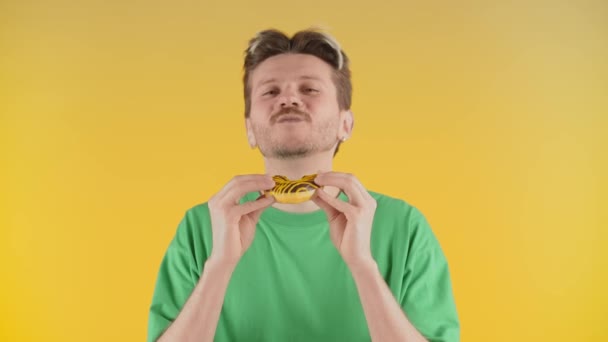 Un giovane con le stoppie gode del sapore di una torta. Un uomo con una maglietta verde mangia una torta in piedi davanti alla telecamera su uno sfondo giallo. Filmati 4k di alta qualità - Filmati, video