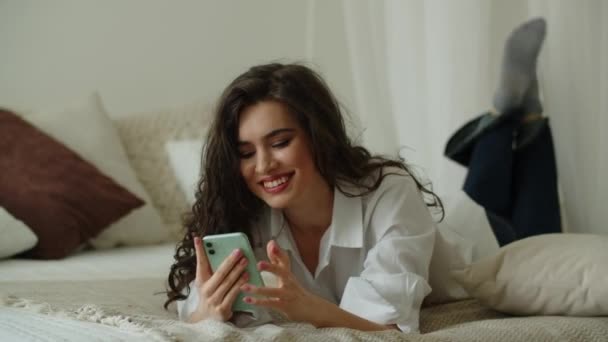 Chica morena usando su teléfono inteligente mientras está acostado en la cama en el dormitorio. Una mujer feliz mandando mensajes a alguien. Imágenes de alta calidad 4k - Metraje, vídeo
