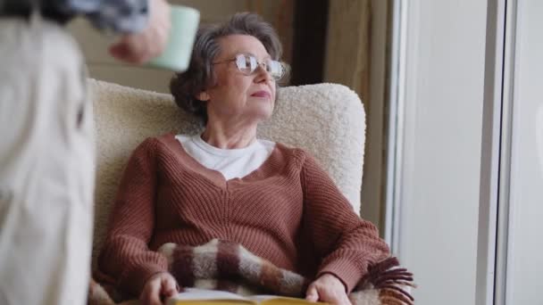 Eine ältere Frau liest in einem gemütlichen Sessel ein Buch, ihr Mann hat ein duftendes Getränk mitgebracht. Hochwertiges 4k Filmmaterial - Filmmaterial, Video