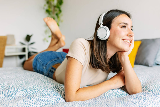 Jeune jolie femme écoutant de la musique avec des écouteurs sans fil tout en étant couché sur le lit. Millennial femelle détente à la maison profiter playlist en ligne. - Photo, image