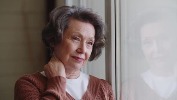 Egy töprengő, elegáns idős hölgy áll egy nagy ablaknál. Emlékek a múltból. Gondolatok az életről. Kiváló minőségű 4k felvételek - Felvétel, videó