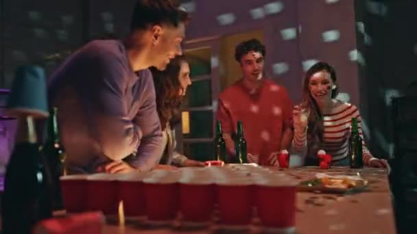 Woman throwing ball in beer cups at table having failed. Excited cheerful friends playing alcohol ping pong at home night party. Happy buddies laughing enjoying girl setback. Carefree weekend hangout. - Footage, Video