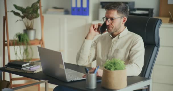 Молодий чоловік-підприємець розмовляє на смартфоні і використовує ноутбук, сидячи за столом в офісі - Кадри, відео