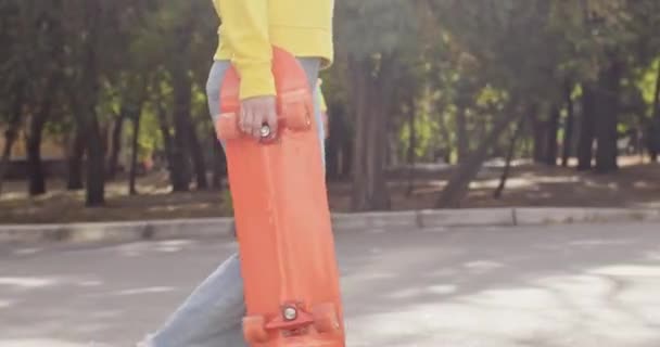 Κορίτσι έφηβος περπατά κατά μήκος πάρκο άσφαλτο δρόμο με skateboard στο χέρι. Κορίτσι βόλτες για να skate park για την πρακτική skateboarding σε ηλιόλουστο καιρό - Πλάνα, βίντεο