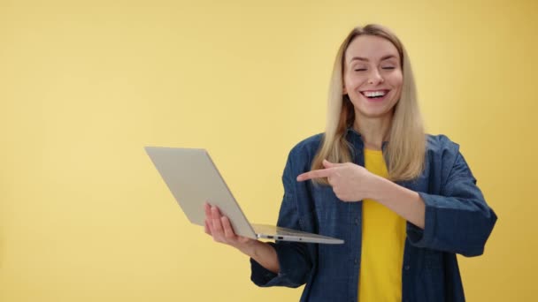 Atraktivní žena ukazující prstem na displej přenosného notebooku, který drží v rukou izolované žluté pozadí studia. Běloška dívající se na kameru s rozpačitým úsměvem uvnitř. - Záběry, video
