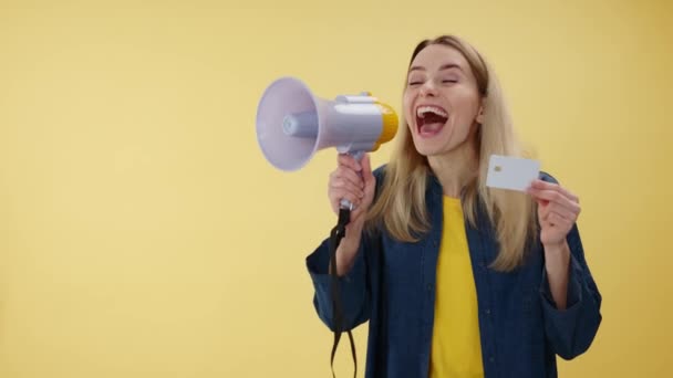 Portrait of crazy female holding credit card and exclaiming in megaphone in studio. Energetic caucasian woman telling about discounts in stores over isolated yellow background. Concept of banking. - Footage, Video
