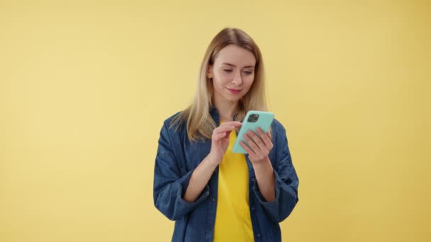 Amazed woman making winner gesture and saying wow while holding modern smartphone in studio. Attactive female looking on device and expressing happy emotions. Isolated over yellow background. - Footage, Video