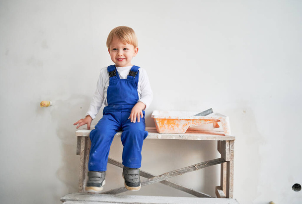 Cute kid construction worker sitting on wooden table against white wall in apartment under renovation. Cheerful little boy wearing safety helmet and work overalls while playing at home. - Photo, Image