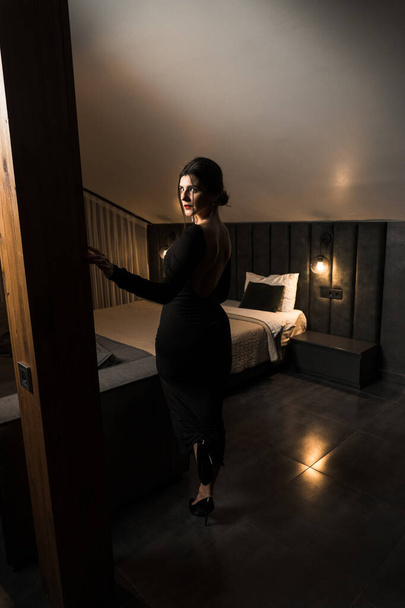 Κομψή γυναίκα με μαύρο φόρεμα που ποζάρει σε διαμέρισμα. Έννοια λήψης μόδας - Φωτογραφία, εικόνα